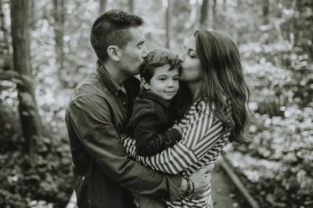 Famille en noir et blanc, les parents embrassant leur fils sur les joues, en forêt
