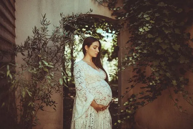 Femme enceinte en robe de dentelle blanche, sous une arche de verdure